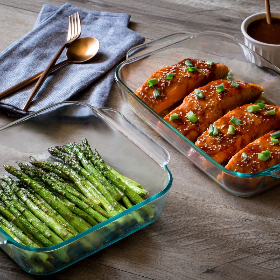 Two glass dishes on a wooden table, one with asparagus and the other with salmon.